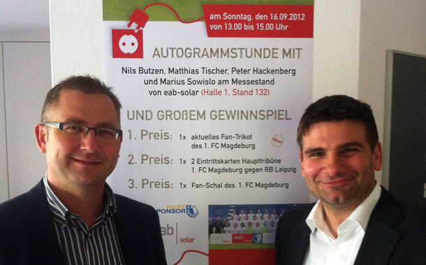 Herr Embach (links) und Herr Hartmayer (rechts) in den Produktionsräumen in Braunschweig vor einem Plakat der EAB Solar mit einer Einladung zu einer Autogrammstunde